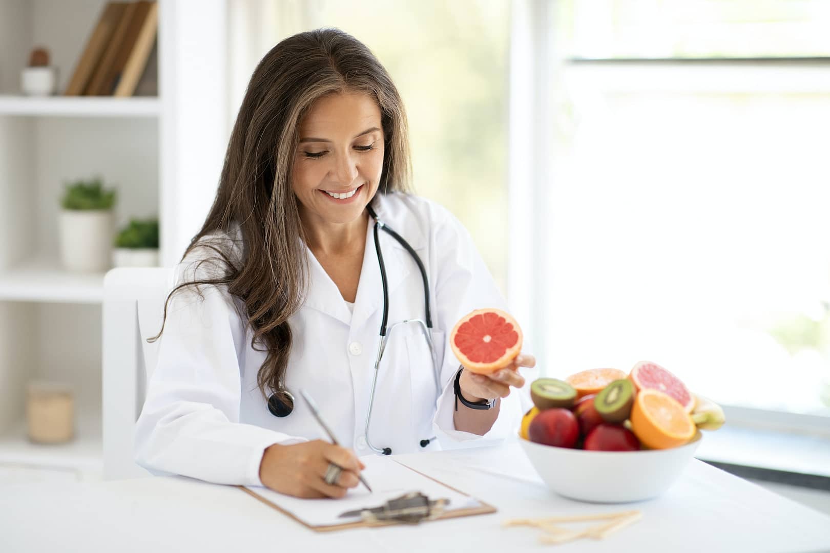 Glad european middle aged woman doctor nutritionist makes notes of diet plan at table with fruits